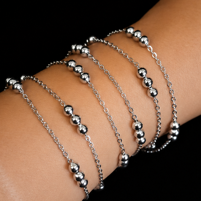 A close-up of a person's wrist wearing a stack of delicate silver chain bracelets, each adorned with small metallic bead clusters, set against a solid black background.
