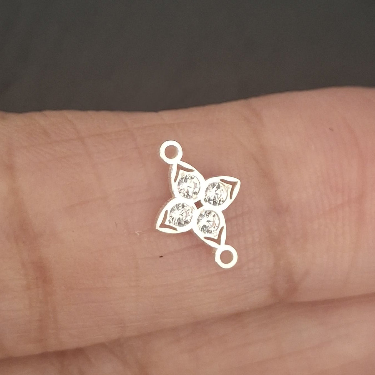 A small silver-toned metal charm or connector with a four-pointed star-like openwork design and loops at the ends, resting on a person's fingers against a dark gray background.