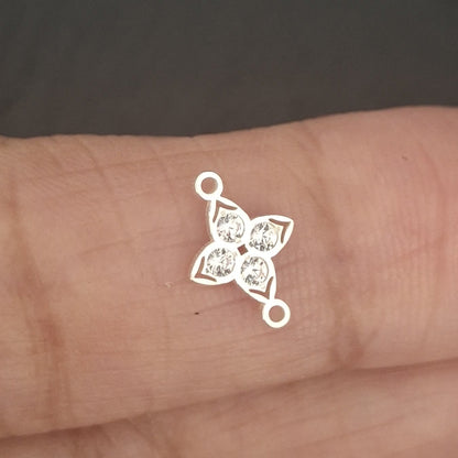 A small silver-toned metal charm or connector with a four-pointed star-like openwork design and loops at the ends, resting on a person's fingers against a dark gray background.